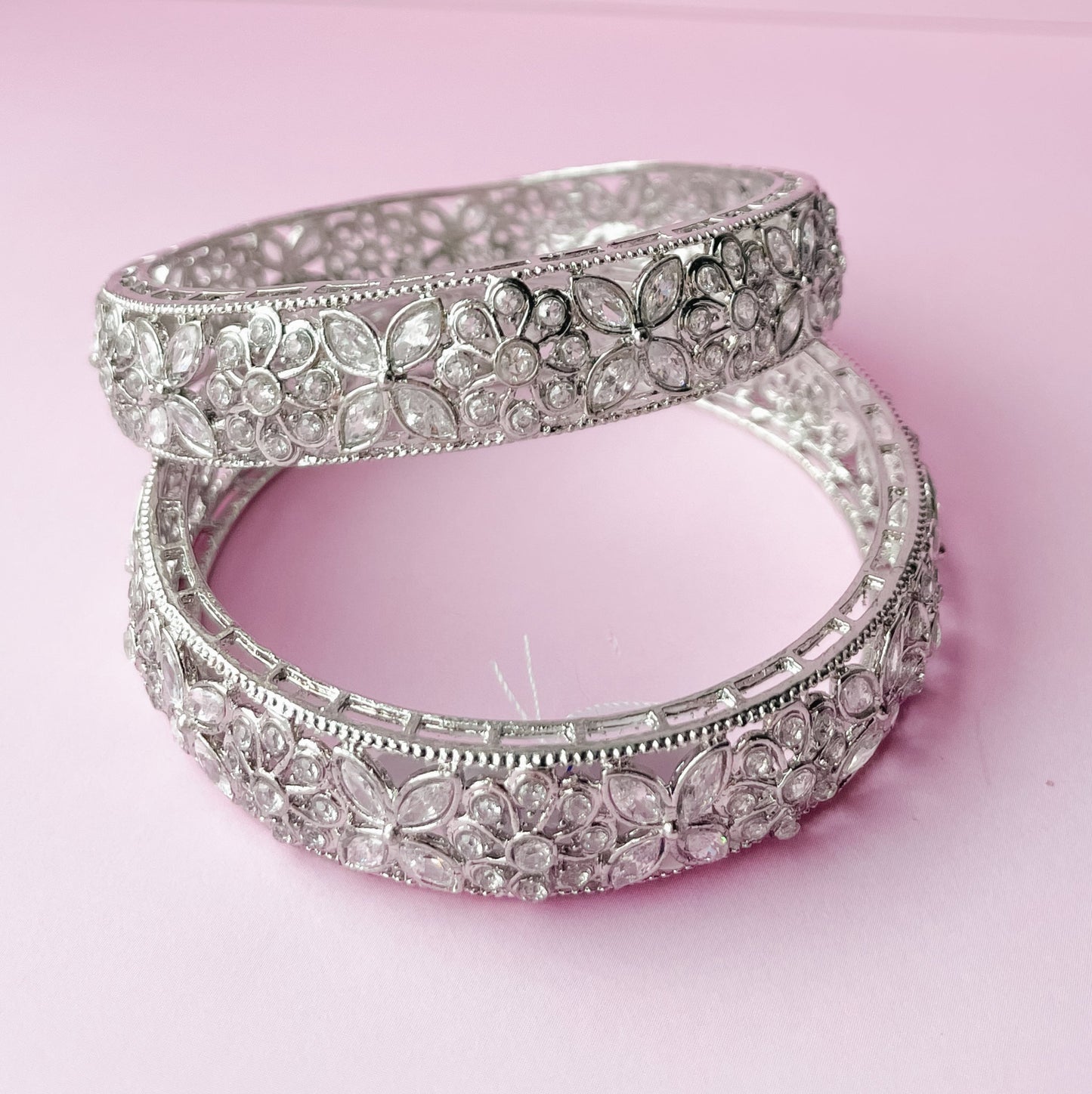 A pair of silver bangle bracelets with floral patterns, displayed on a pink background.