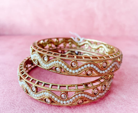 Two gold-colored bangle bracelets with floral patterns and adorned with pearls displayed on a pink background.