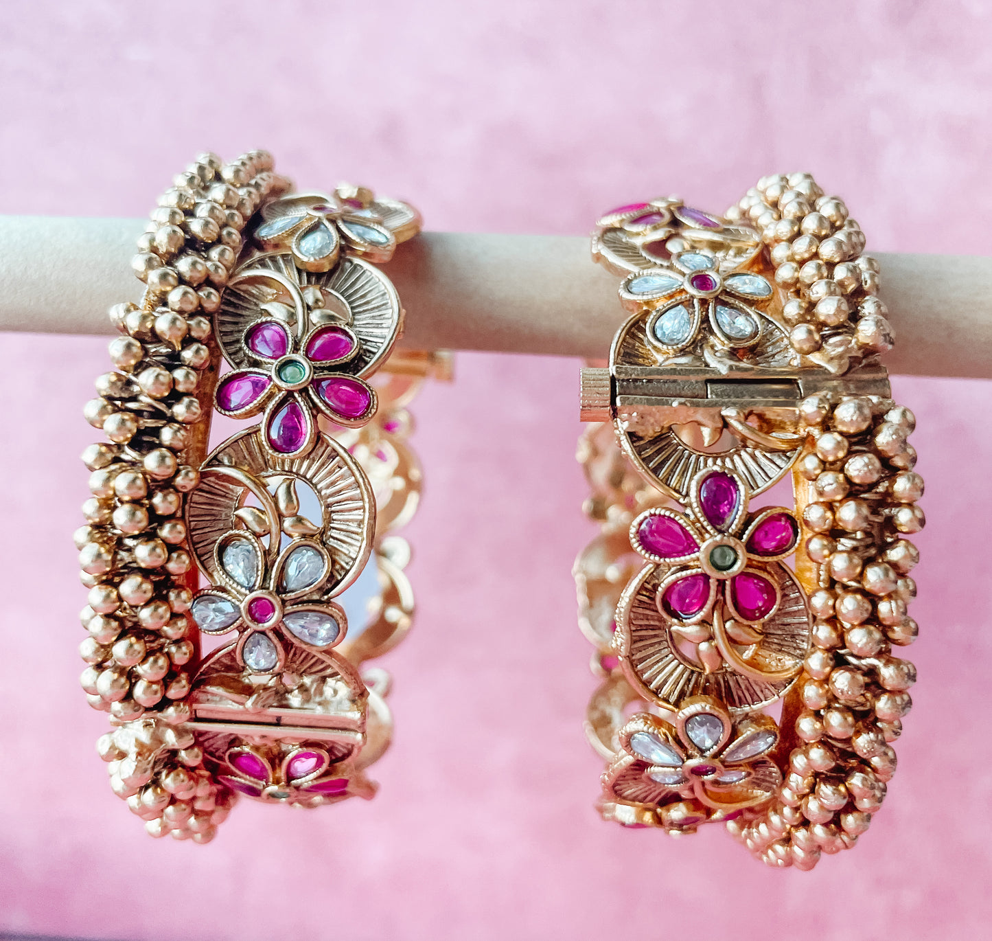 Two gold-colored bracelets with floral patterns and adorned with pink and red beads and pearls displayed against a pink background.