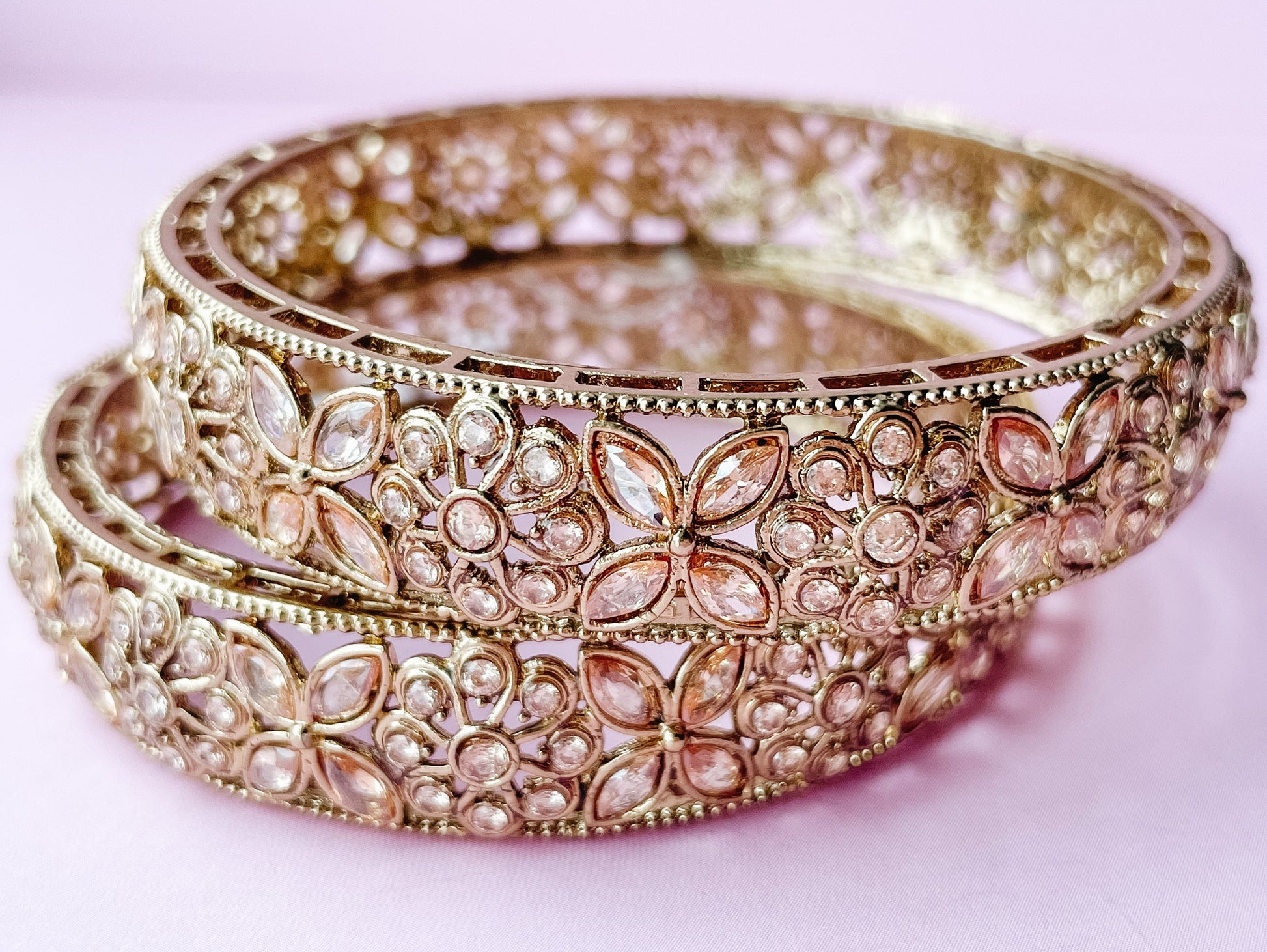 Two gold bangles with intricate designs on a light pink background