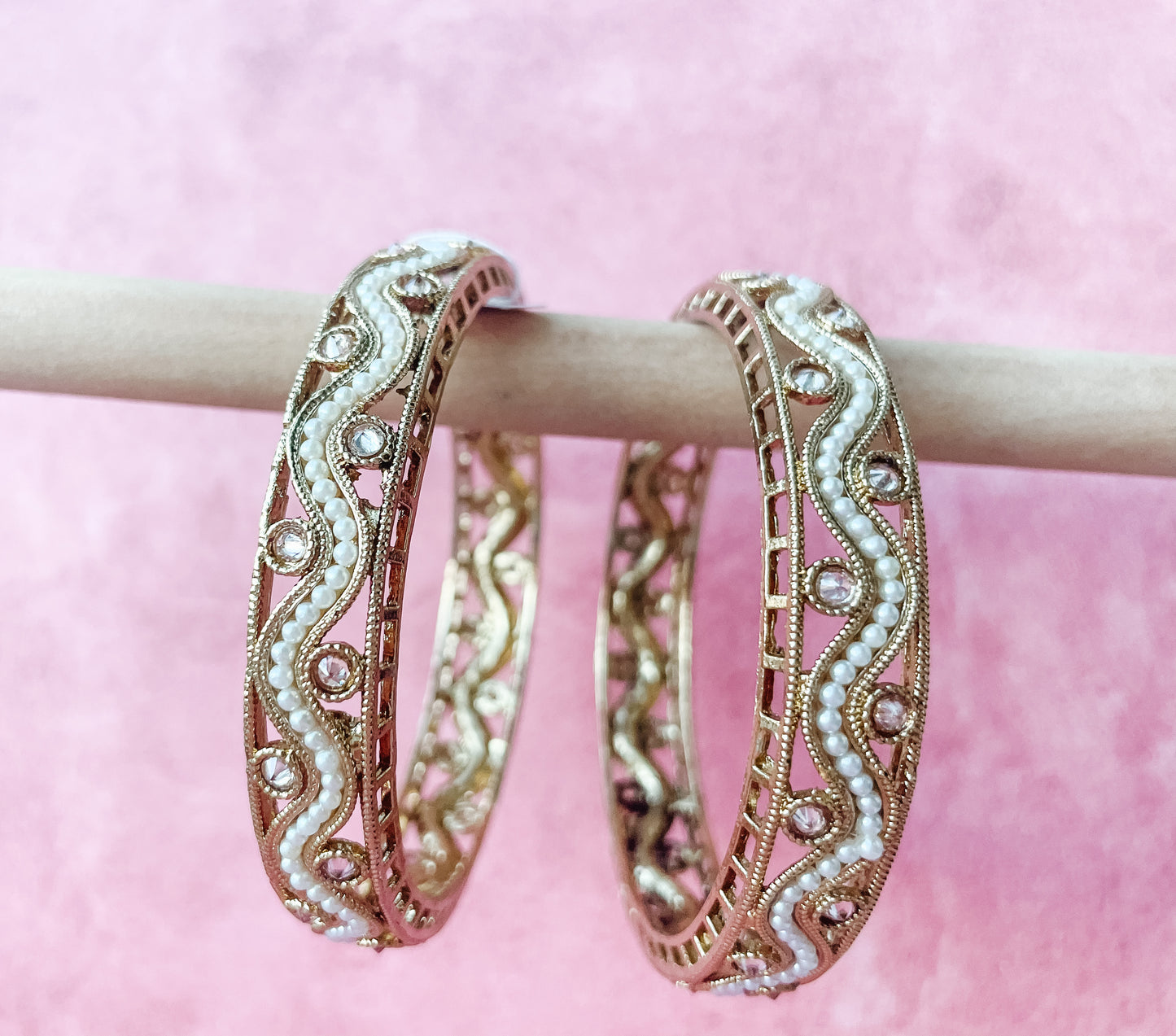 Two gold-colored bangle bracelets with floral patterns and adorned with pearls displayed on a pink background.